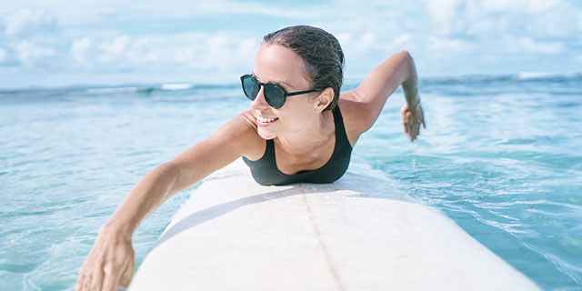 Woman on surfboard with sunglasses on, paddling in water. Skybright Colloidal Silver Aloe Vera Gel – essential for New Zealand Summer.